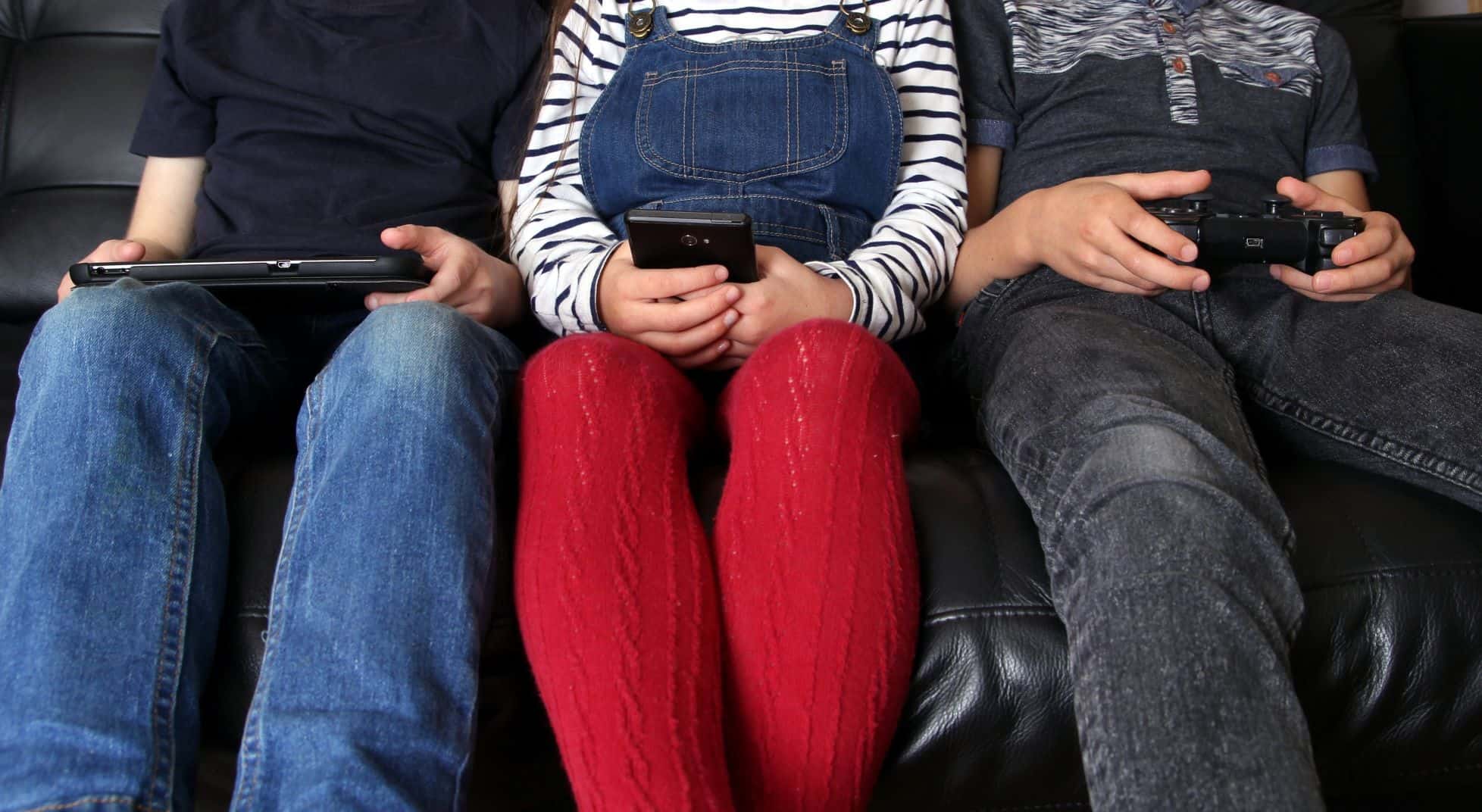 Three young adults each using a device--a tablet, a cell phone, and a remote controller.
